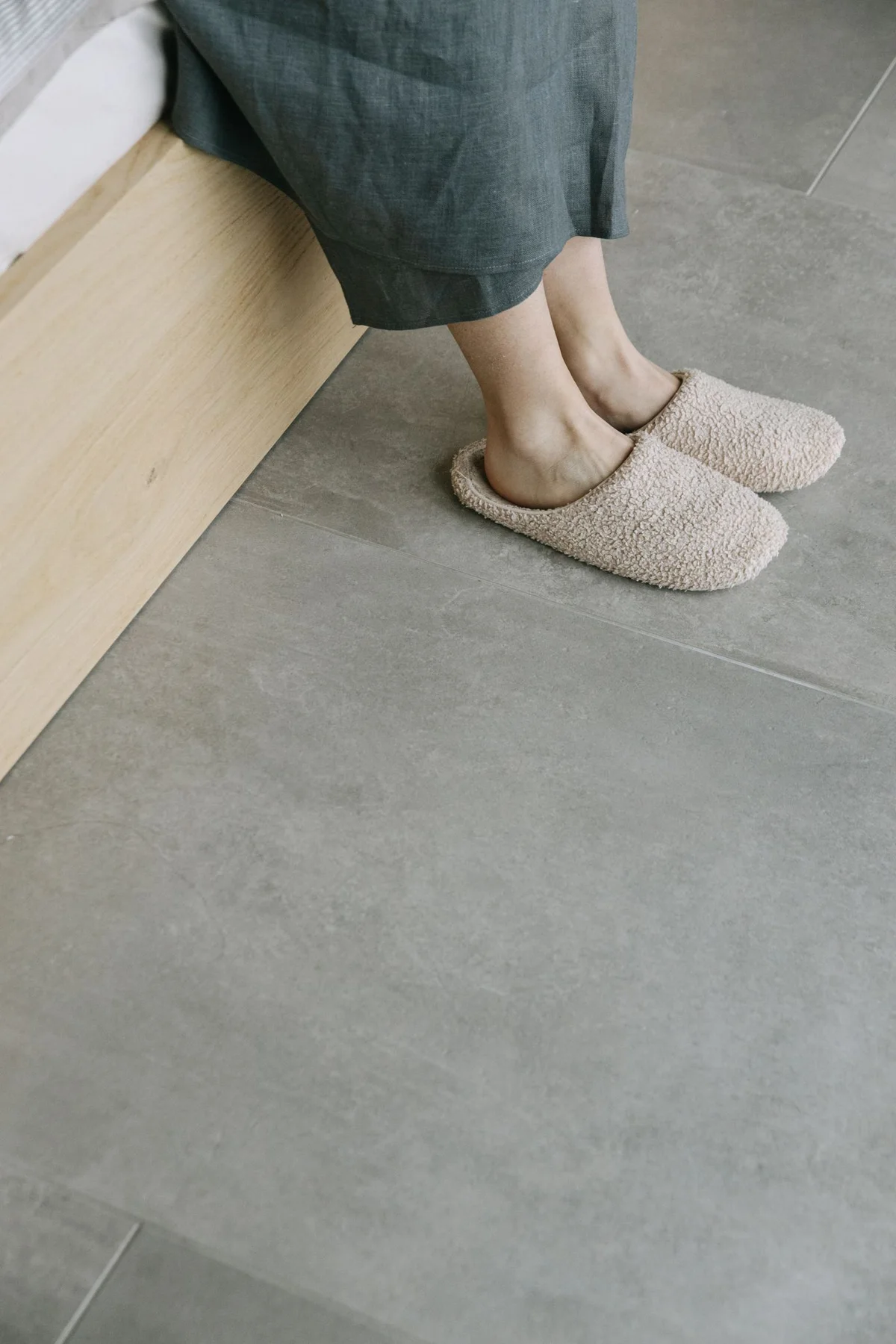 A close view of feet in indoor slippers beside a bed.