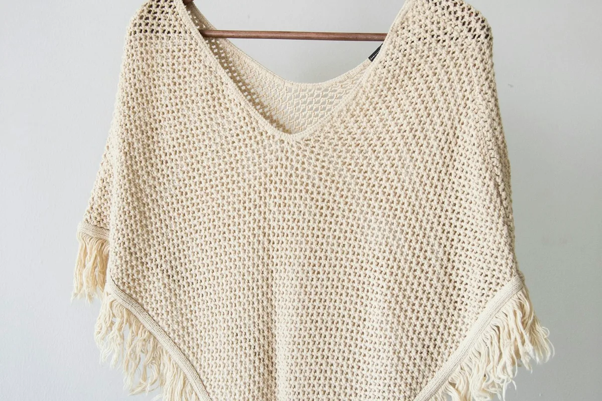 A pale open-textured knitted garment with fringed edges on a hanger.