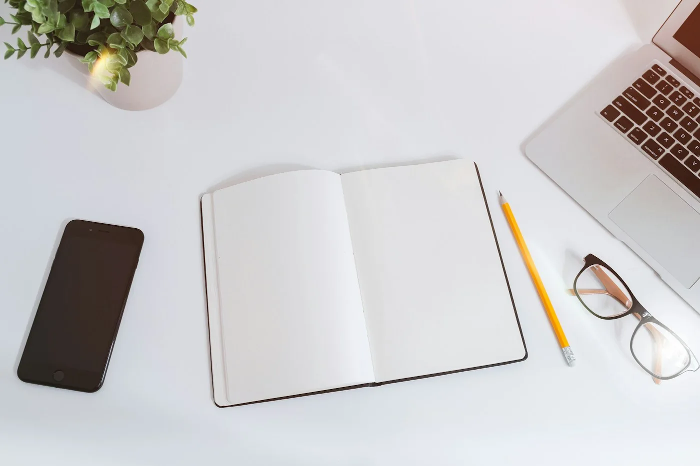 An open notebook, pencil, glasses, phone and laptop on a pale desk.